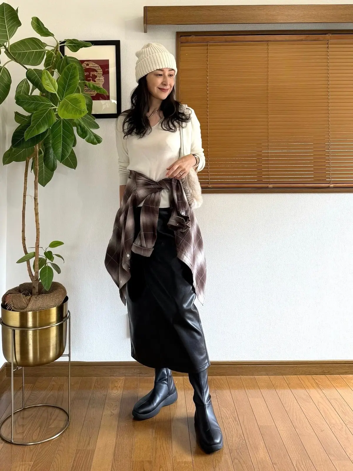 白ニットに黒のレザーロングスカート、黒いニーハイブーツを合わせた女性。首には赤茶色のチェック柄シャツを巻き付け、頭には白のニット帽を被っている。背景には観葉植物と木製のブラインド。
GUフェイクレザースカートのコーディネート。