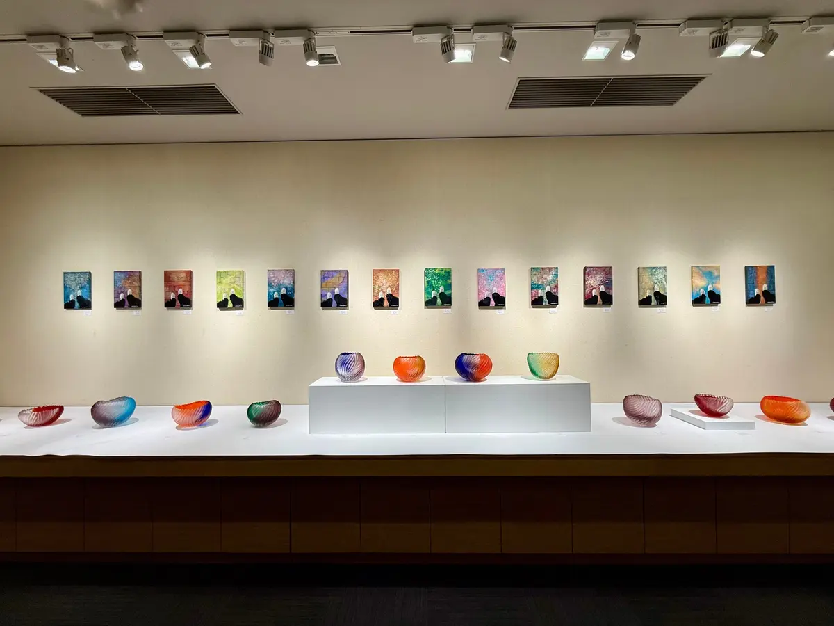 美術館の展示室。白い壁に18枚の絵画が並び、下にはガラス製のオブジェが置かれた展示台。天井にはスポットライトが設置されている。土田康彦　僕が僕であるために　あべのハルカス　大阪　ヴェネツィアンガラス