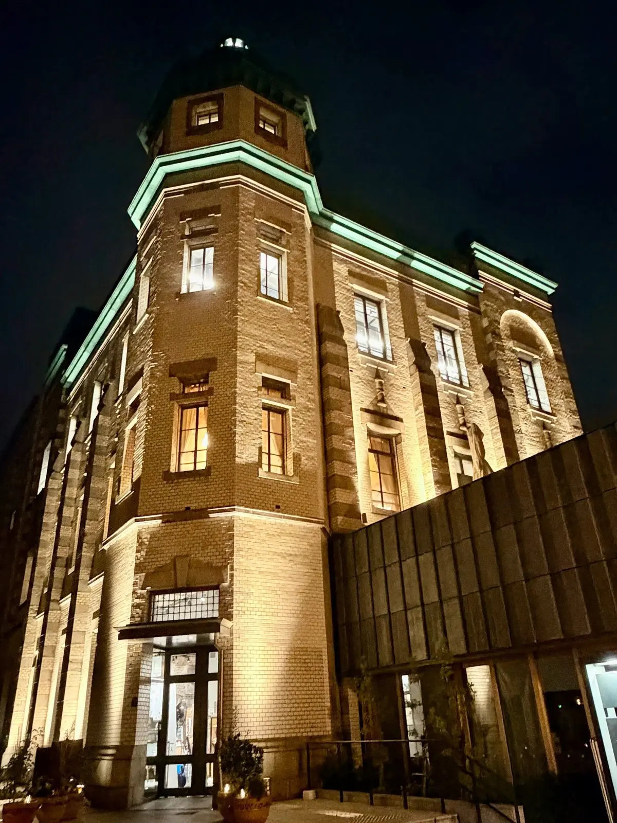 埼玉県川越市りそなコエドテラス。夜の暗闇に浮かぶ、レンガ造りの多層式建物。右側に現代的な金属製の壁面があり、左側には隣接する建物の一部が見える。建物には複数の窓があり、一部が光っている。角には緑色の装飾が施された高い塔がある。