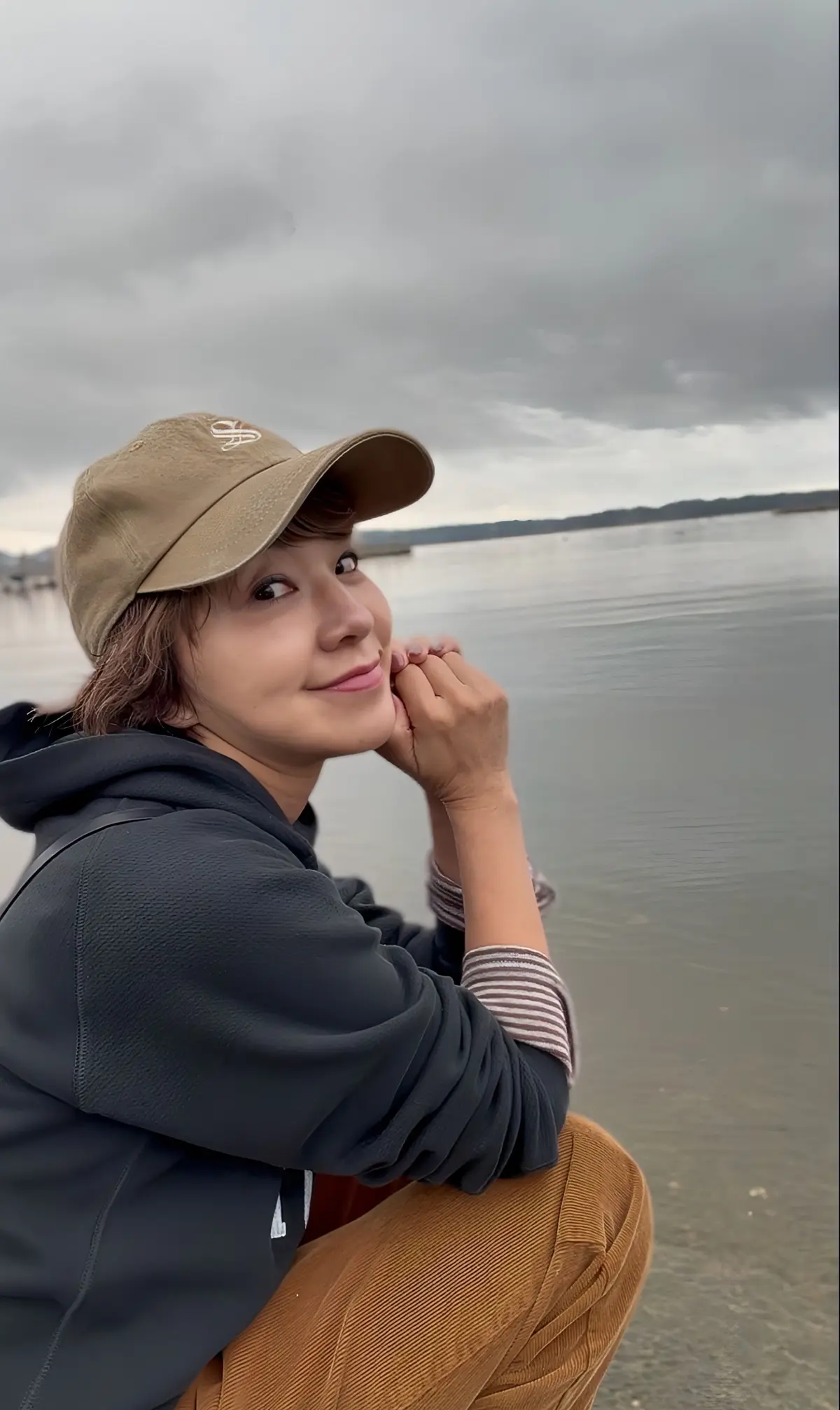 ベージュのキャップを被った女性が、水辺に座ってこちらを見ている。グレーのパーカーを着ており、右手を顎に当てている。背景は曇り空の海。