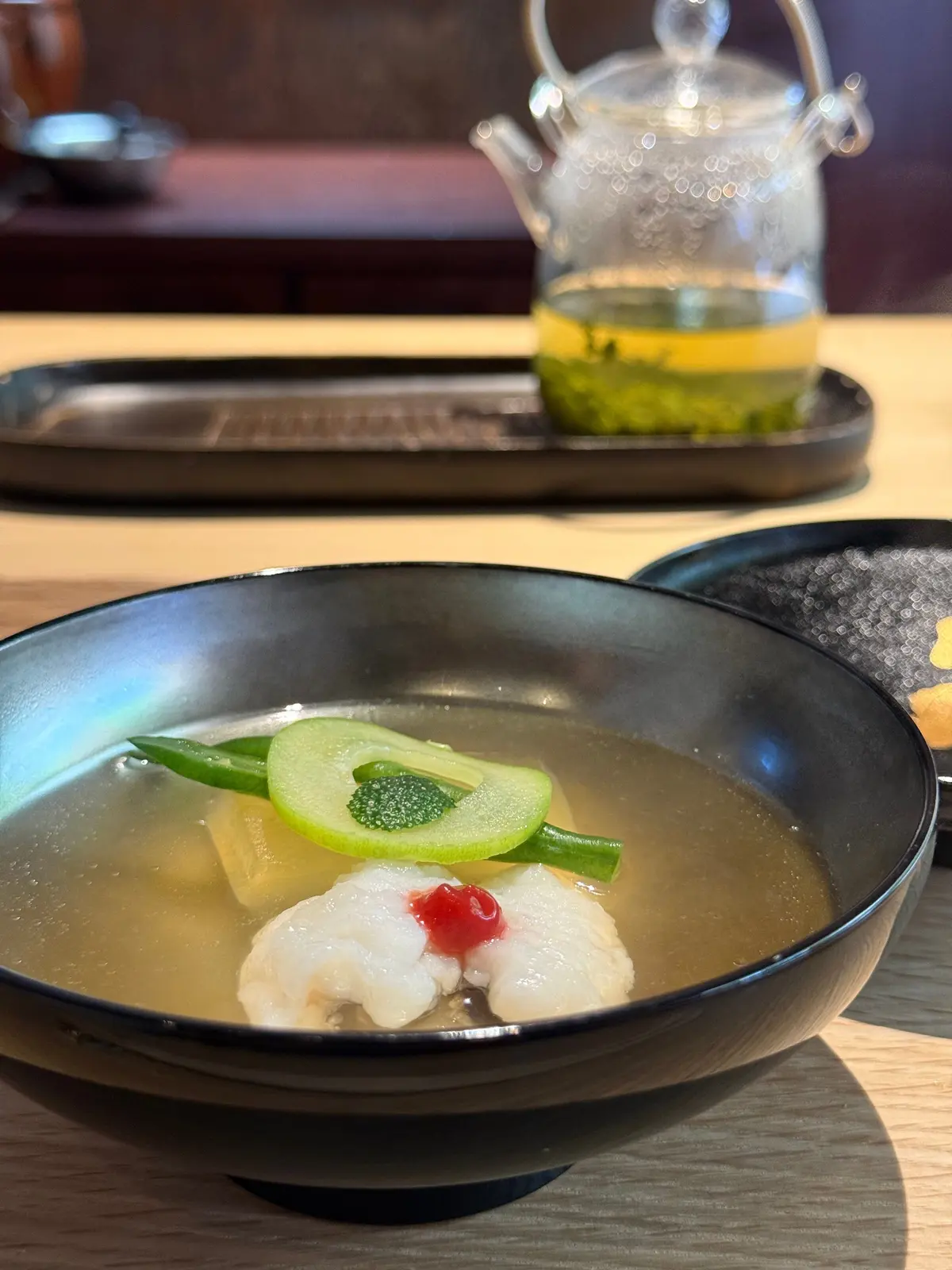 黒い漆器の椀に、透明な澄んだスープ。白身魚、キュウリ、グリーンの葉、赤い実が盛り付けられている。背景には、緑色の葉が入ったガラスの急須。