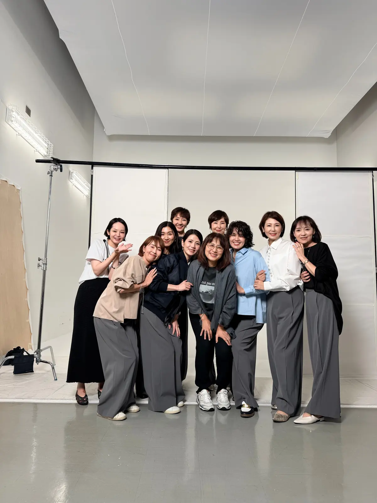 白い壁のスタジオで、グレーのパンツスーツやカジュアルな服装を着た10人の女性が笑顔で写っている。手前に7人、奥に3人が並んでおり、一部の人々は手を振っている。撮影機材が左側に設置されている。