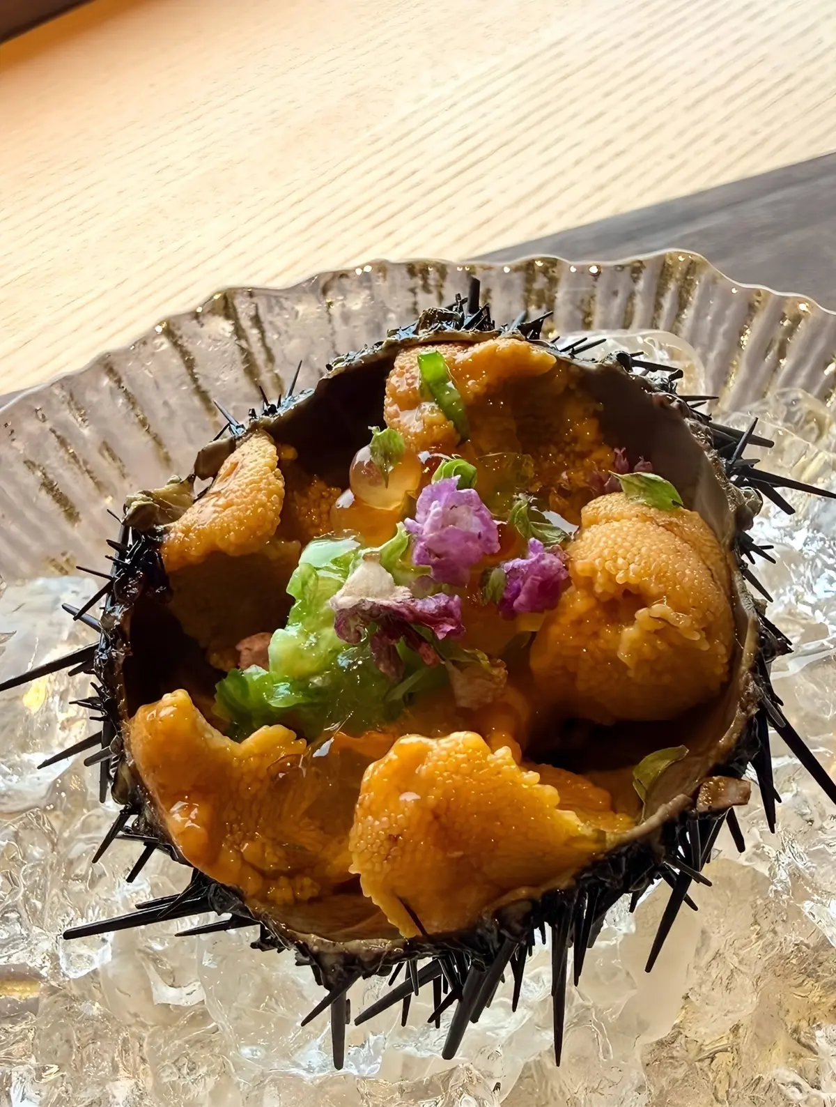 真珠のような鮮やかなオレンジ色のウニが、黒いトゲの殻に盛り付けられた料理。彩り豊かな葉野菜や紫色の花が添えられている。