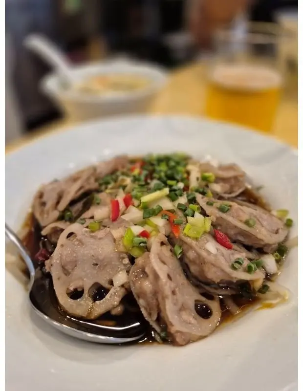 白磁の皿に盛り付けられた、薄切りにした豚肉とレンコンの冷菜。照り気のある濃い色のタレと、赤や緑の細切り野菜が散らされている。背景には、スープとビールが置かれた木製のテーブル。