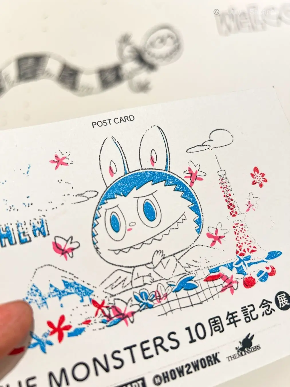 青いウサギのキャラクターが手紙を書く様子を描いたポストカードで、背景に東京タワーと花が描かれている