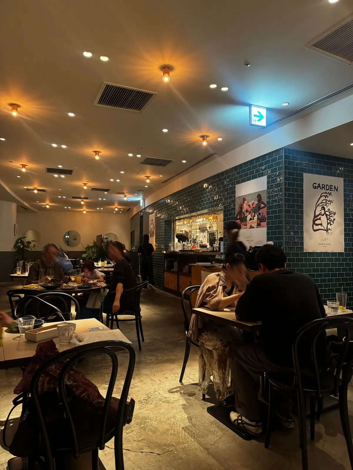 レストランの店内。テーブル席に座って食事をする客が数組。奥にはカウンターがあり、壁面は青色のタイルで装飾されている。天井にはスポットライトが設置され、温かい光が室内を照らしている。右壁には「GARDEN ROSE」と書かれたポスターが貼られている。