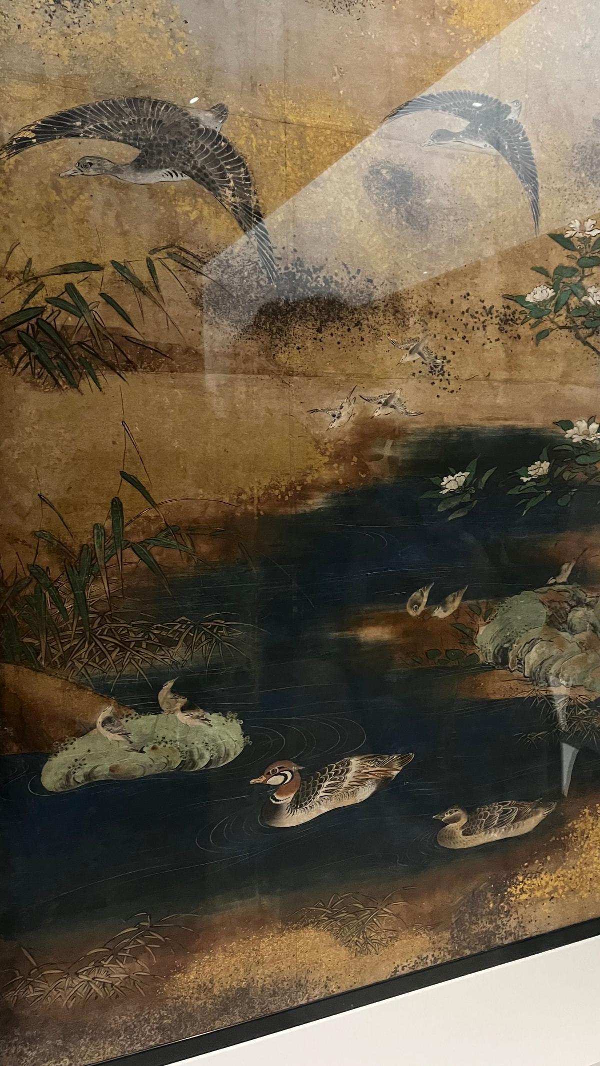 金色の背景に、水辺を飛ぶカモや水鳥、葦や蓮の花が描かれた水墨画風の絵画。上部には大きな鳥が舞い、下部にはカモの親子が泳いでいる。