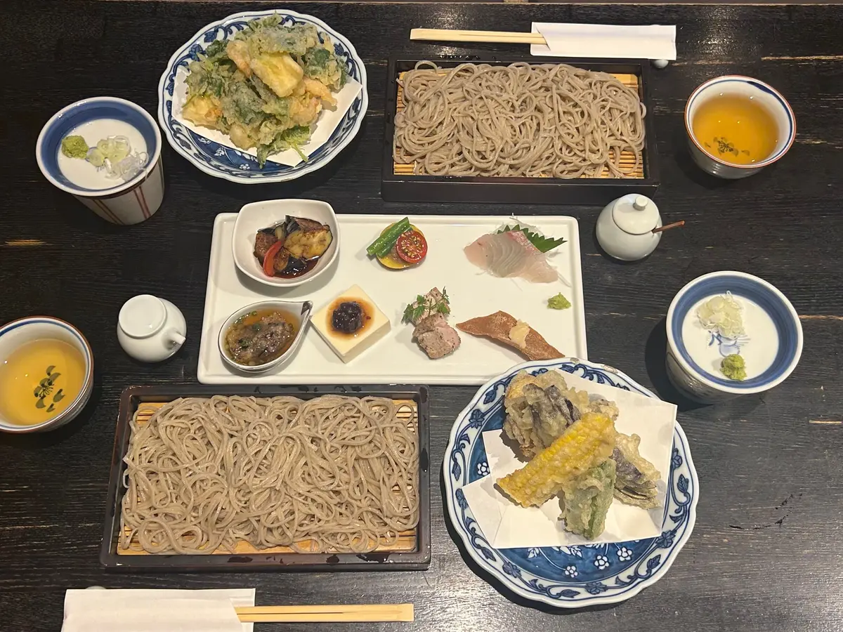 木製のテーブルに並べられた、蕎麦の盛り合わせ。蕎麦、天ぷら、だし汁、小鉢などが、藍色の器や木製の盆に盛り付けられている。