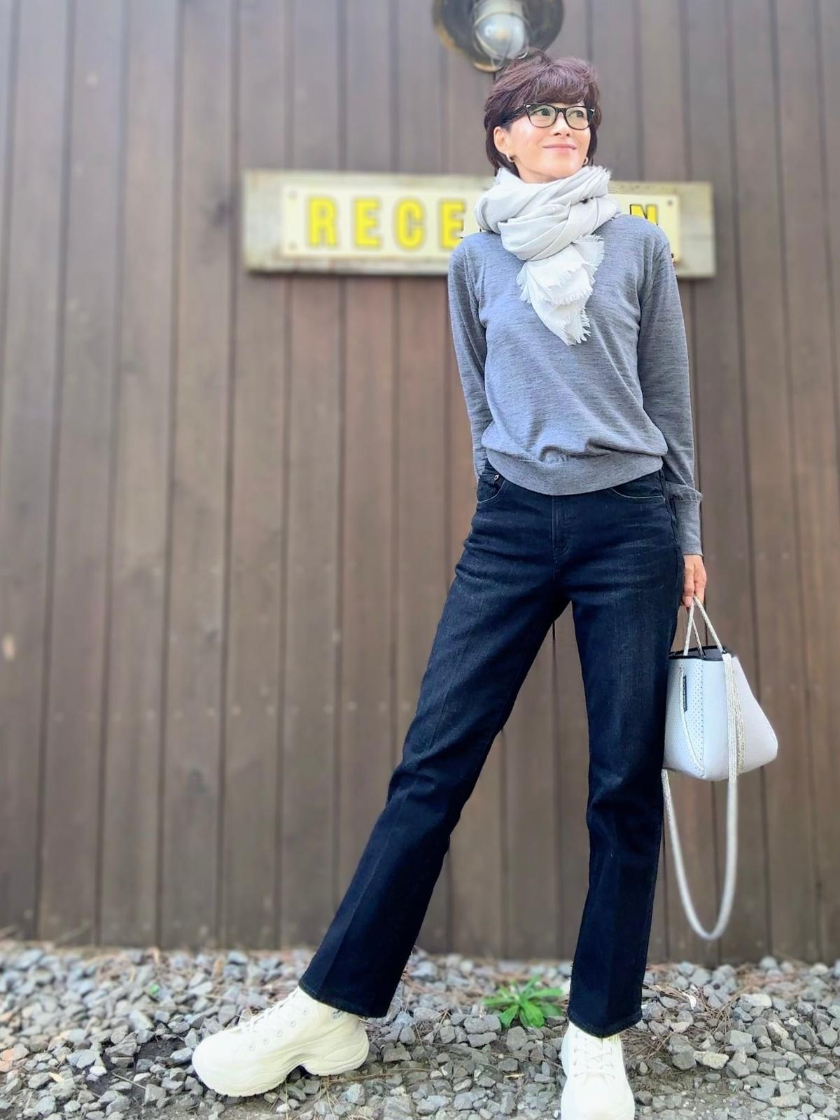 グレーのセーターにデニム、白いスニーカーを合わせた女性。白いスカーフを着用し、ライトブルーのハンドバッグを持つ。背景は木製の壁。