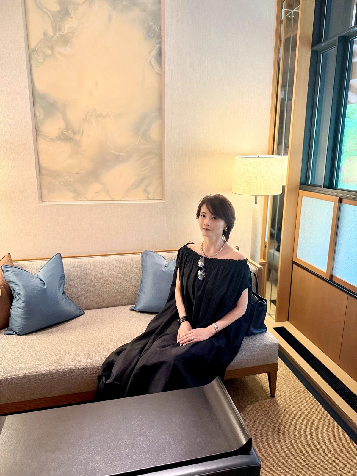 黒いオフショルダーのワンピースを着た女性が、ベージュのソファに座っている。背景には抽象画と間接照明のランプが置かれている。