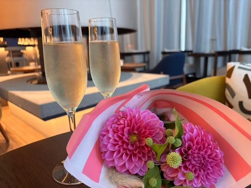 テーブルに置かれたシャンパングラス2杯とピンクと白のラッピングされたピンクのダリアの花束。背景にはレストランのテーブル席と窓が見える。