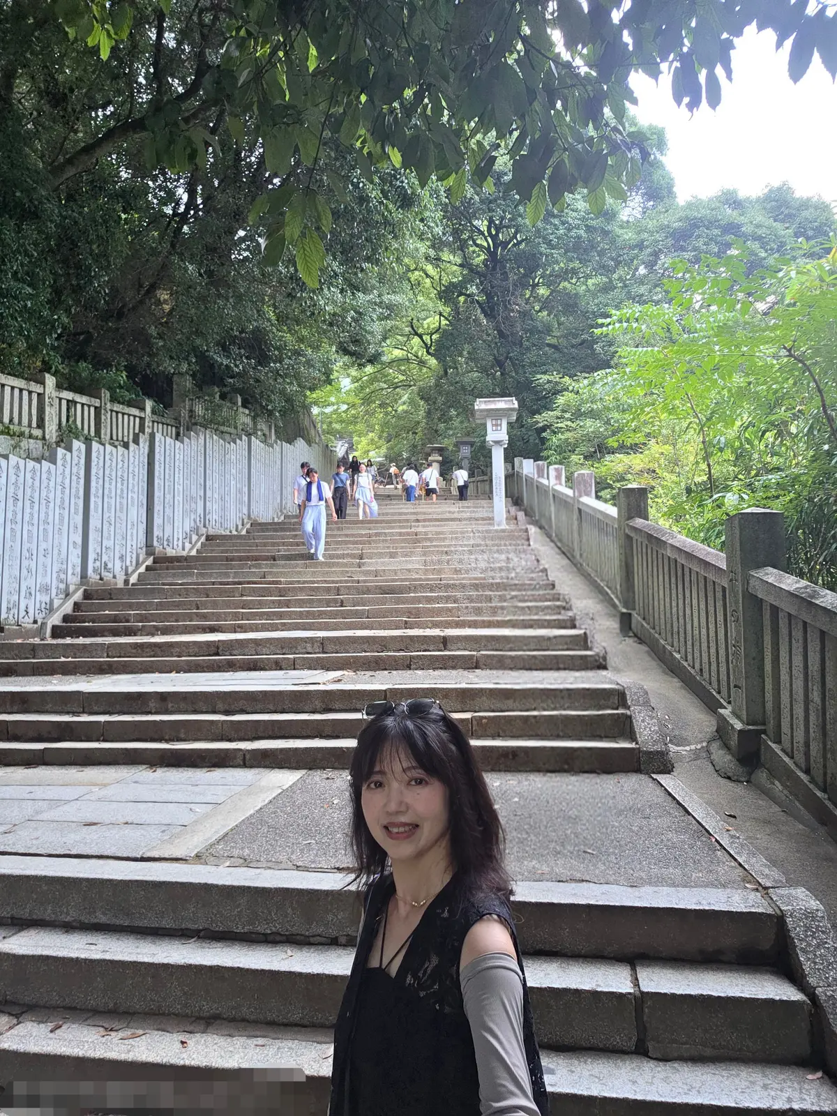石段の上で微笑む女性。緑豊かな木々が茂る背景に、多くの人が階段を上っている様子が見える。