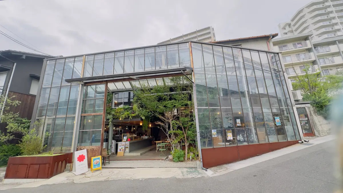 ガラス張りのカフェ storefront、緑の植物と木製の家具が並ぶ。建物の周りには高層ビルが立ち並んでいる。