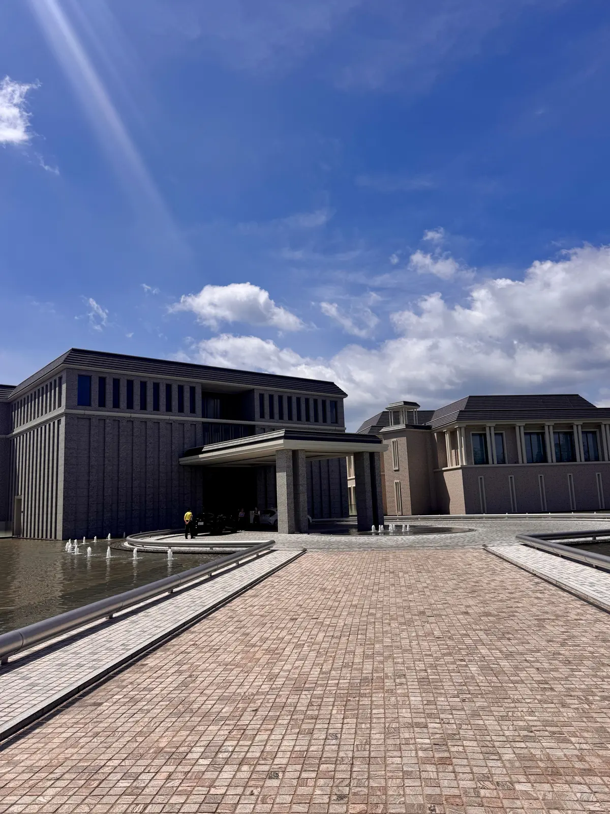 現代的なデザインの建物と水庭園。グレーの石材で覆われた建物は、直線的な屋根と列柱を持つ。建物前には曲線を描く水辺があり、噴水が設置されている。広場は石畳で舗装され、中央には円形の装飾がある。青空を背景に、洗練された雰囲気が伝わる。