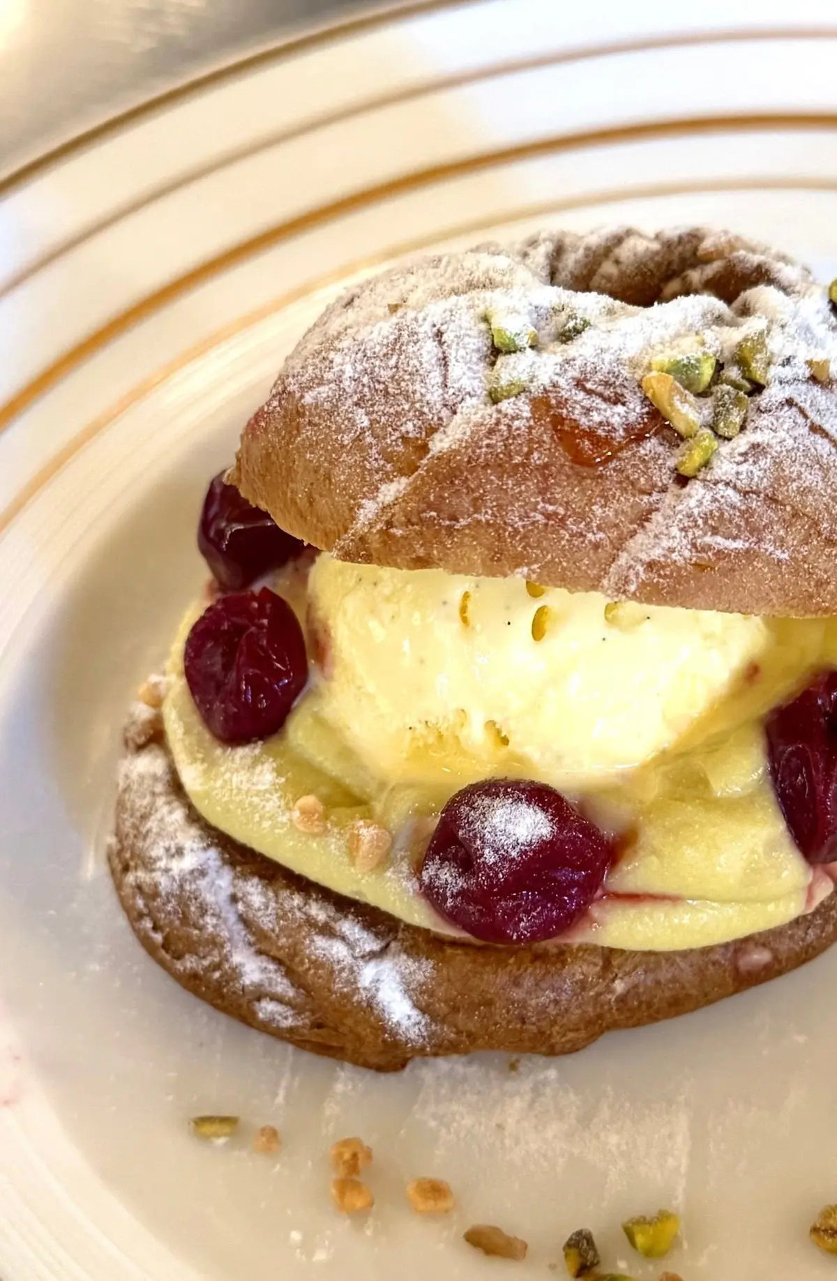 粉砂糖をかけたシュー生地に、バニラアイスクリームとダークチェリー、ピスタチオが添えられたデザート。淡い黄色の皿に盛り付けられている。