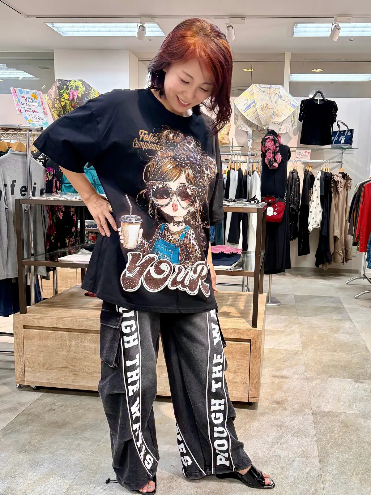 赤い髪の女性が、黒いカーゴパンツと、誕生日のお祝いメッセージとイラストがプリントされた黒色のTシャツを着ている全身写真。背景には店舗の商品棚が見える。