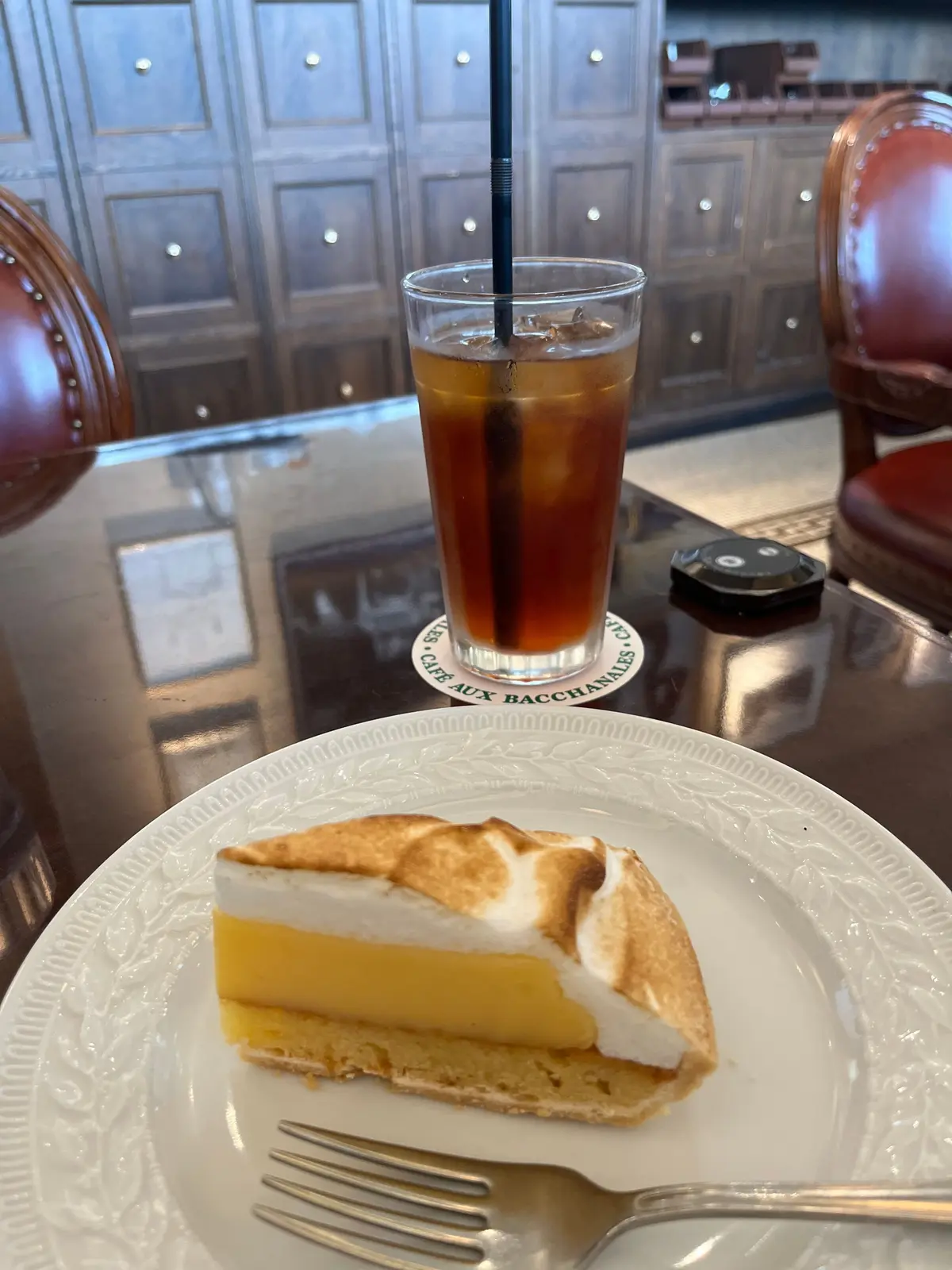 カフェ・オ・バッカナルのコースターの上に置かれた、レモンパイとアイスコーヒー。レモンパイは三角形にカットされ、ホイップクリームと焦げたマシュマロがトッピングされている。背景には木製のテーブルと椅子。