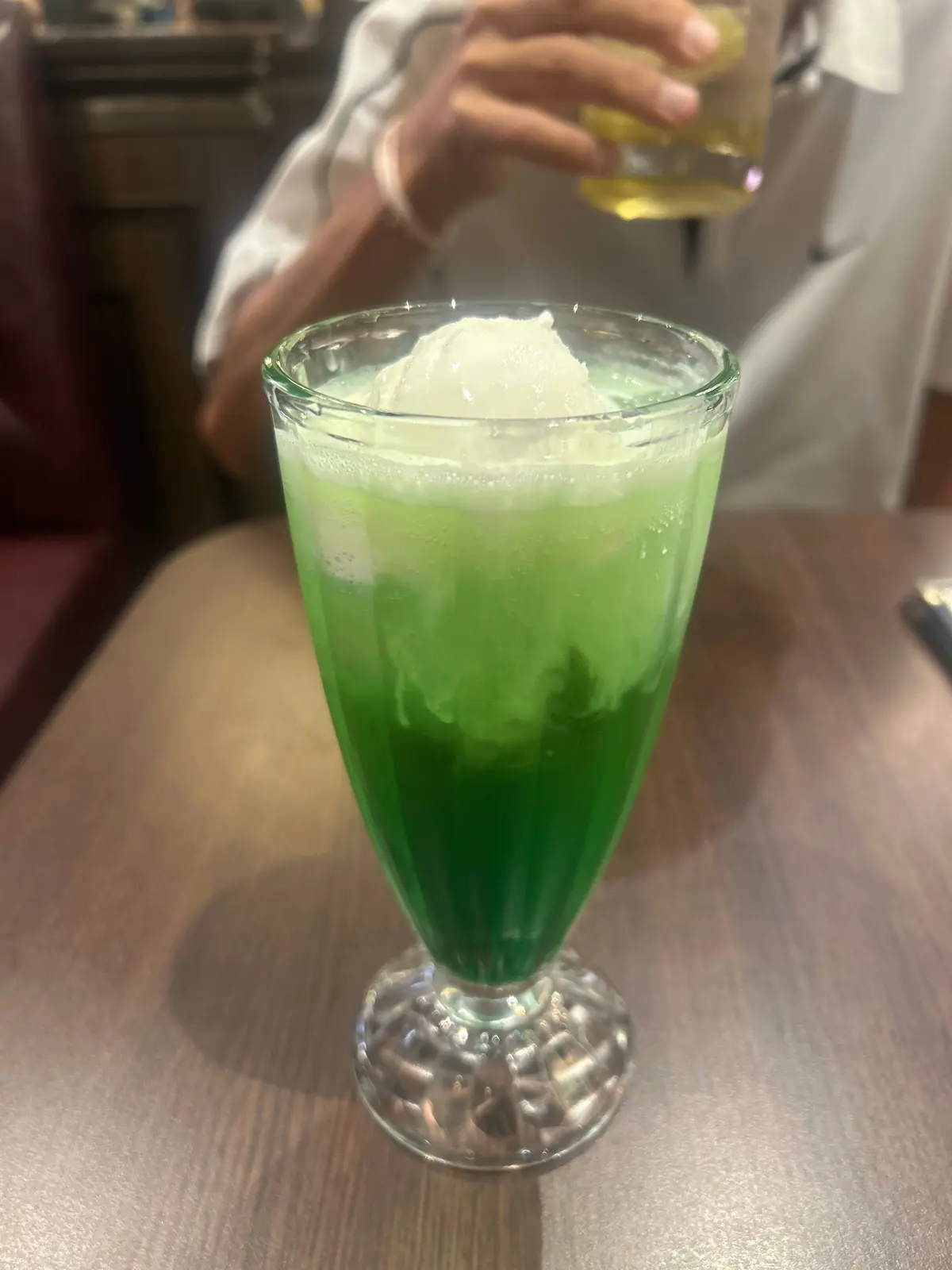 緑色の飲み物が、脚付きのグラスに入っている。氷がたっぷり、上にクリームが乗っている。背景には、白い服を着た人の手と、別の飲み物が写っている。