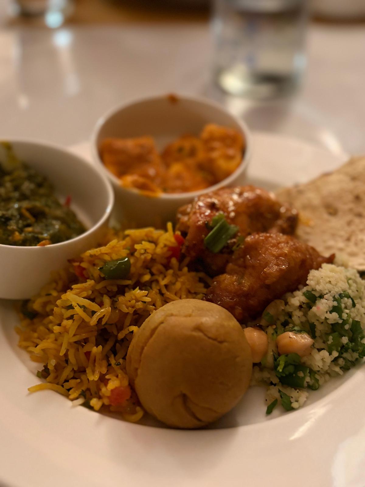 白いお皿に盛り付けられたインド料理。左上にはほうれん草のカレー、右上にはオレンジ色のチキンカレー、中央には黄色いピラフとフライドチキン、右下には野菜とナッツの和え物、手前にはパンが置かれている。