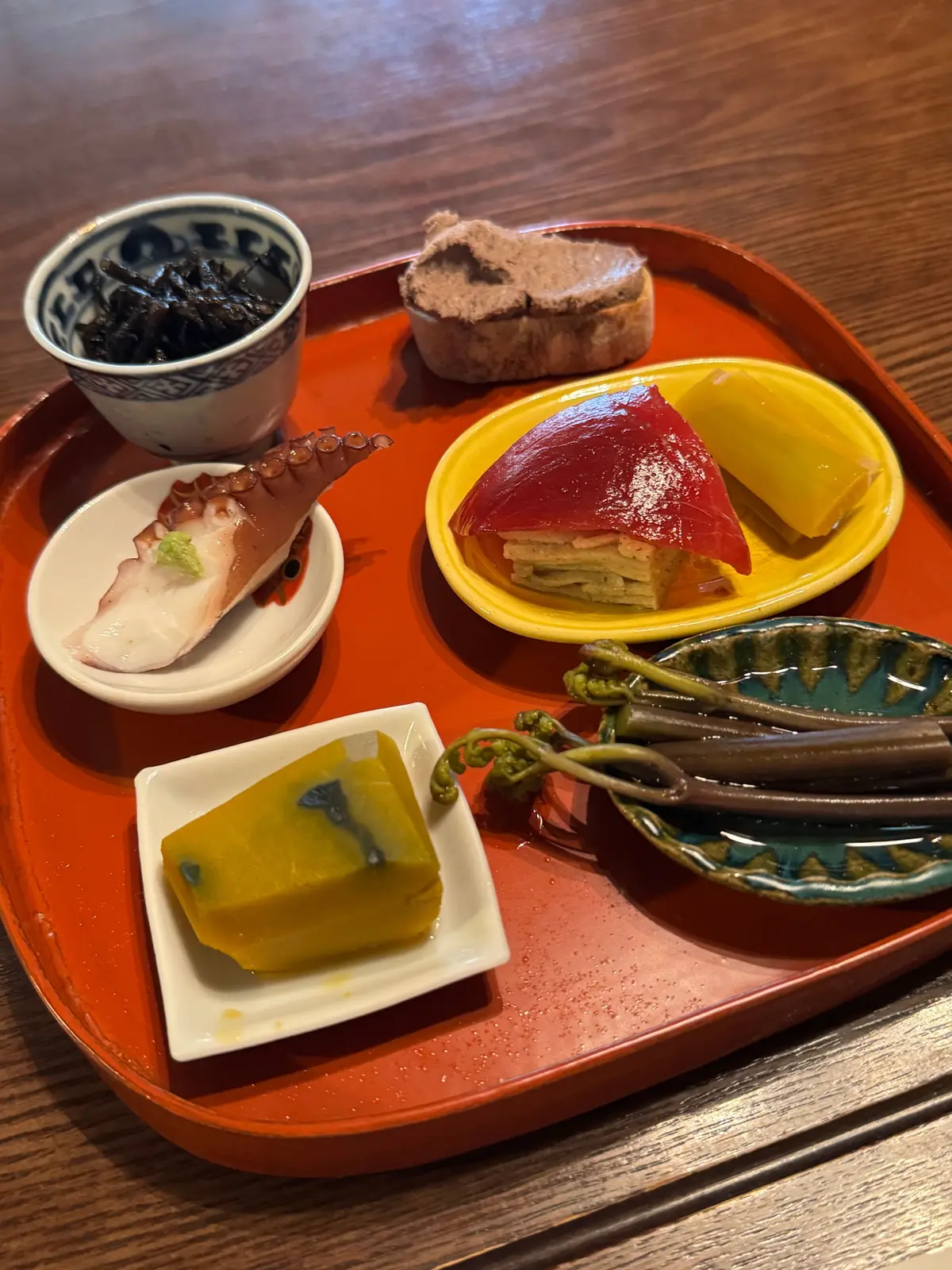 赤色の漆塗りの木製プレートに、様々な日本料理の小鉢が盛り付けられている。黒い海苔、茶色い豆腐、赤いかぶの漬物、ピンク色の魚介類、緑色のゼリー、黄色と青色のゼリー、そして緑色の野菜の小鉢が含まれる。