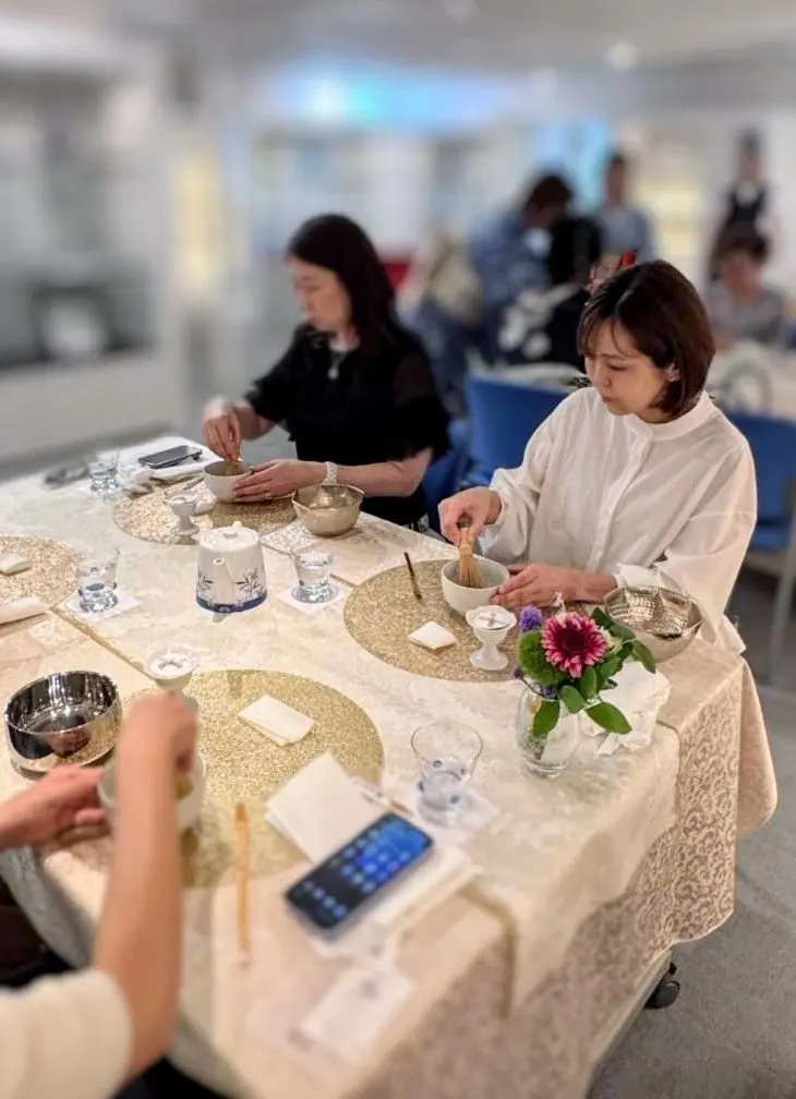 テーブルを囲んで、女性たちが茶碗に粉を入れながら作業している様子。白いテーブルクロスが敷かれ、各席にグラスや花瓶が置かれている。背景には他の人々がぼやけて写っている。