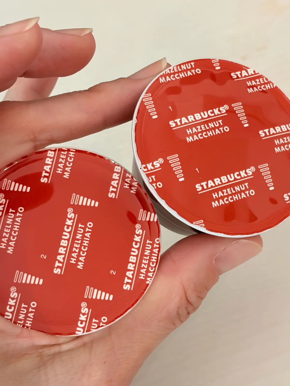 赤色の丸いポッドが複数、指でつまんで立体的に配置されている。ポッドには「STARBUCKS HAZELNUT MACCHIATO」の文字が印刷されている。