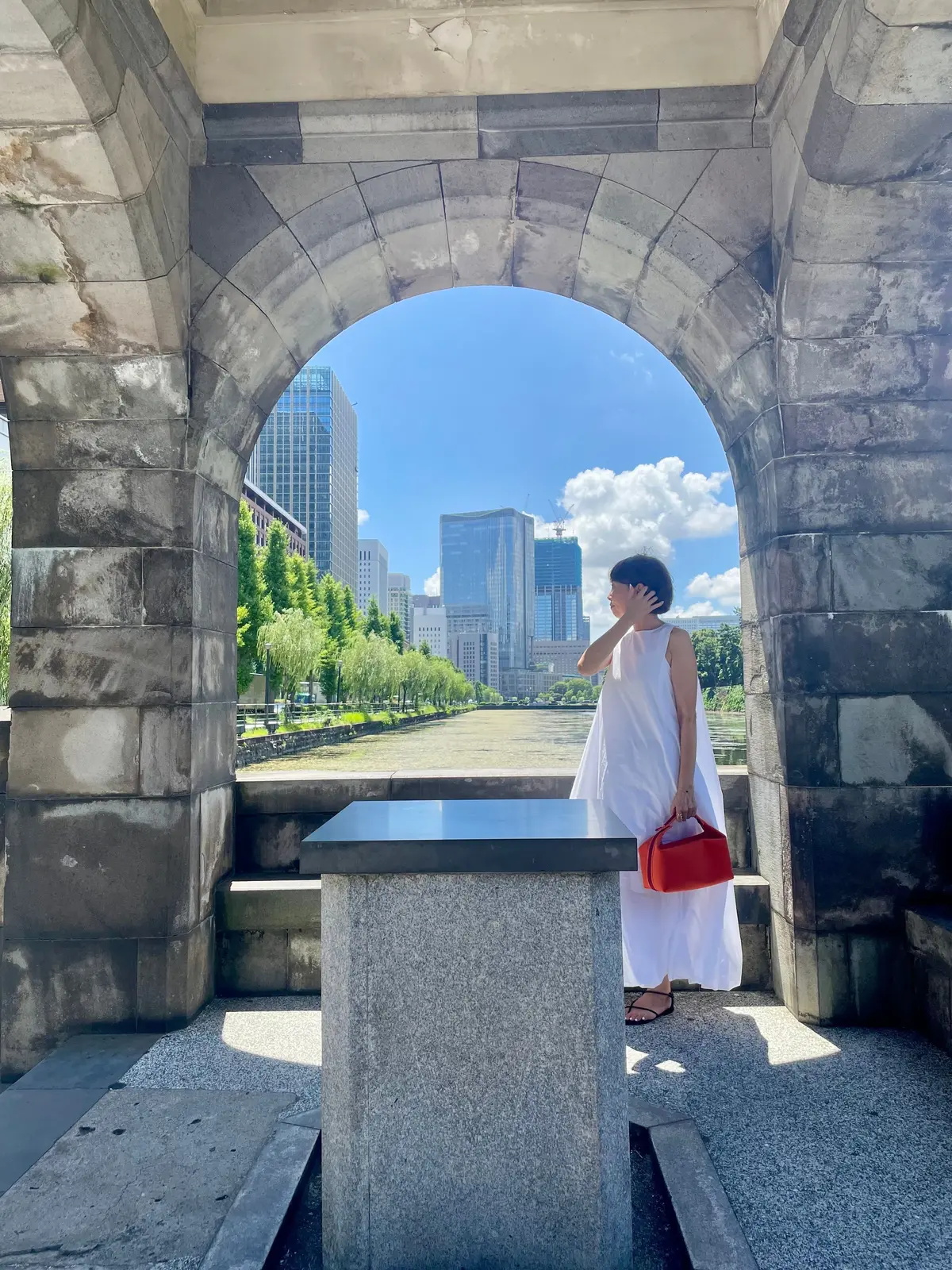 白いワンピースを着た女性が、石造りのアーチを背景に立っている。アーチの向こうには、水辺と高層ビル群が広がり、晴天の空が広がっている。女性は赤いハンドバッグを持ち、こちらを見ている。