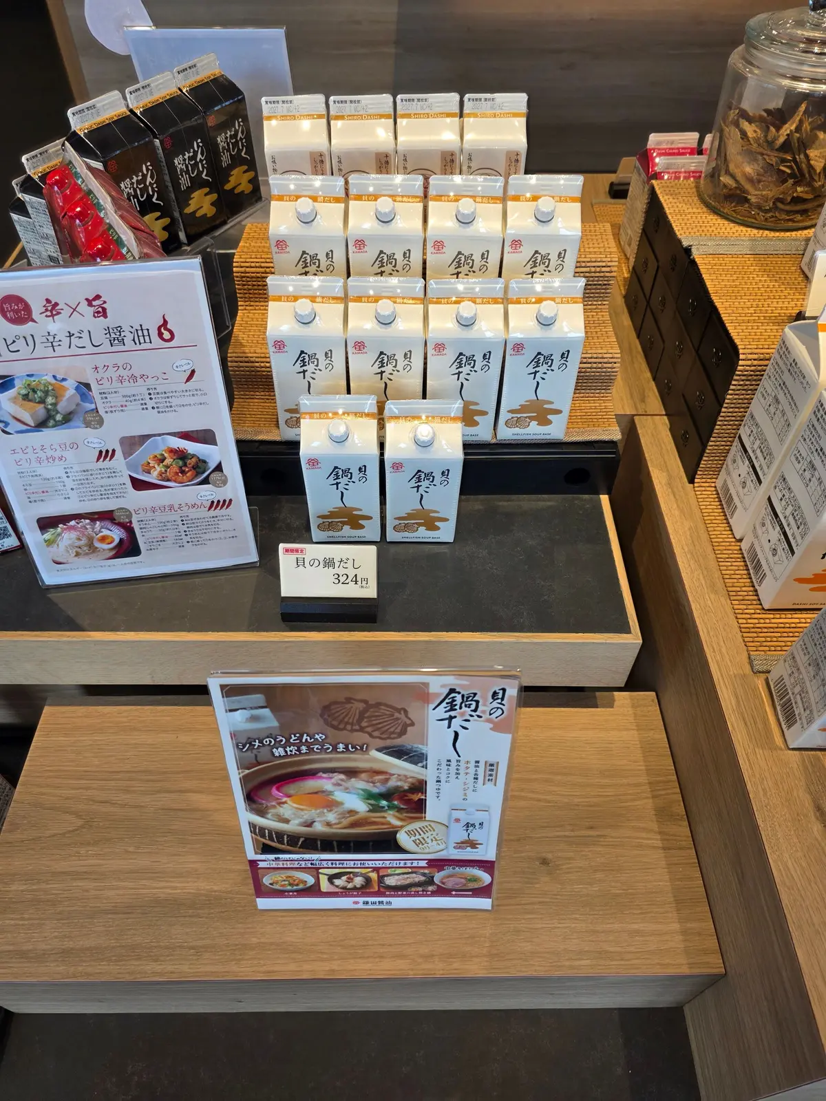 木製のテーブルに、様々な調味料や食品が並んでいる。手前には「鶏旨」と書かれた調味料のボトルが整然と並び、その下にメニューのようなものが置かれている。背景には、他の商品やパッケージが見える。