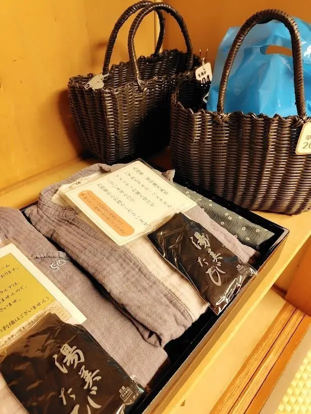 茶色の編みかご2つと、グレーと黒のTシャツが並んだ木製のトレイ。価格表と説明文が添えられ、青いビニール袋も見える。