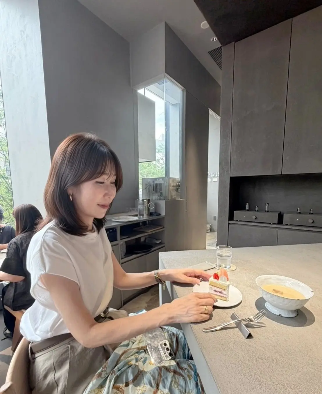 カフェでケーキと飲み物を楽しむ女性。白いTシャツにベージュのスカートを着用し、短めの茶髪ボブ。窓際に座り、ケーキを手に取って見つめている。背景にはグレーの壁とキッチン設備が見える。