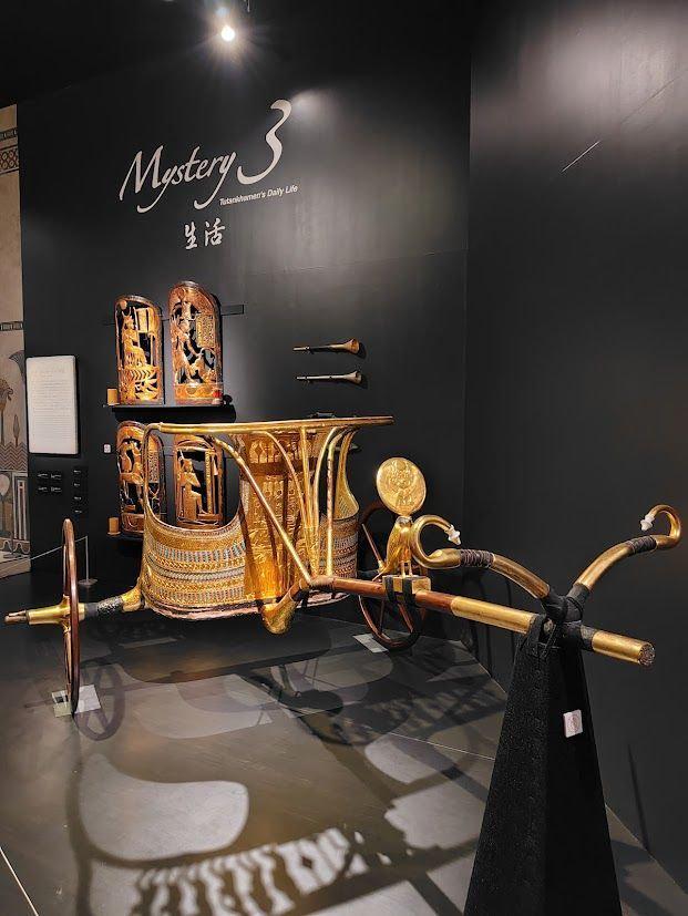 古代エジプトの王の乗車用リヤン車と楽器の展示。金色のリヤン車は装飾が施され、背後には楽器が置かれている。黒い背景に「Mystery 3」の文字が浮かび上がる。