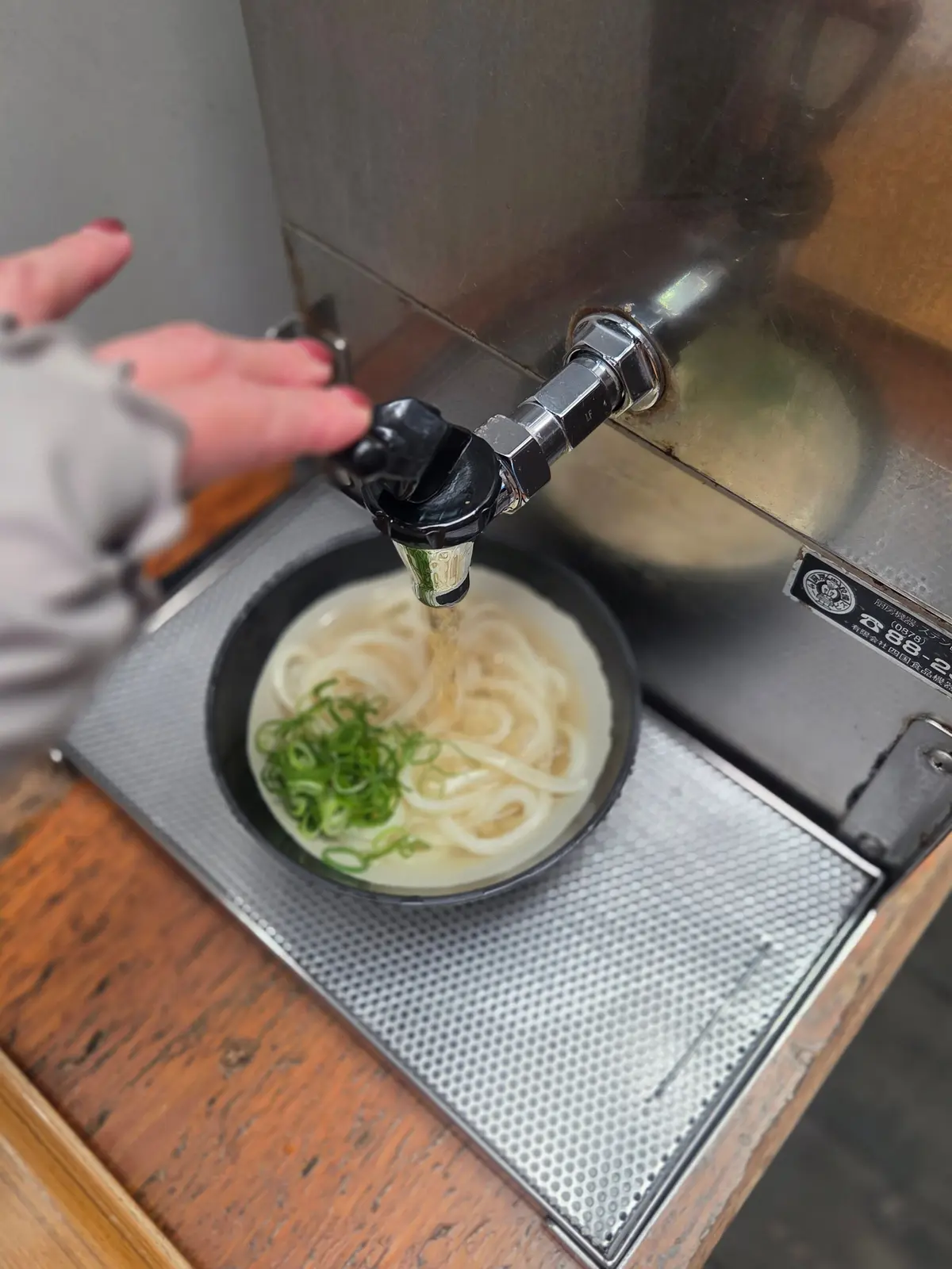 温かい麺料理が黒い皿に入れられ、緑のネギが添えられている。左手で蛇口からお湯が注がれている。背景には金属製の機械と木製の台がある。