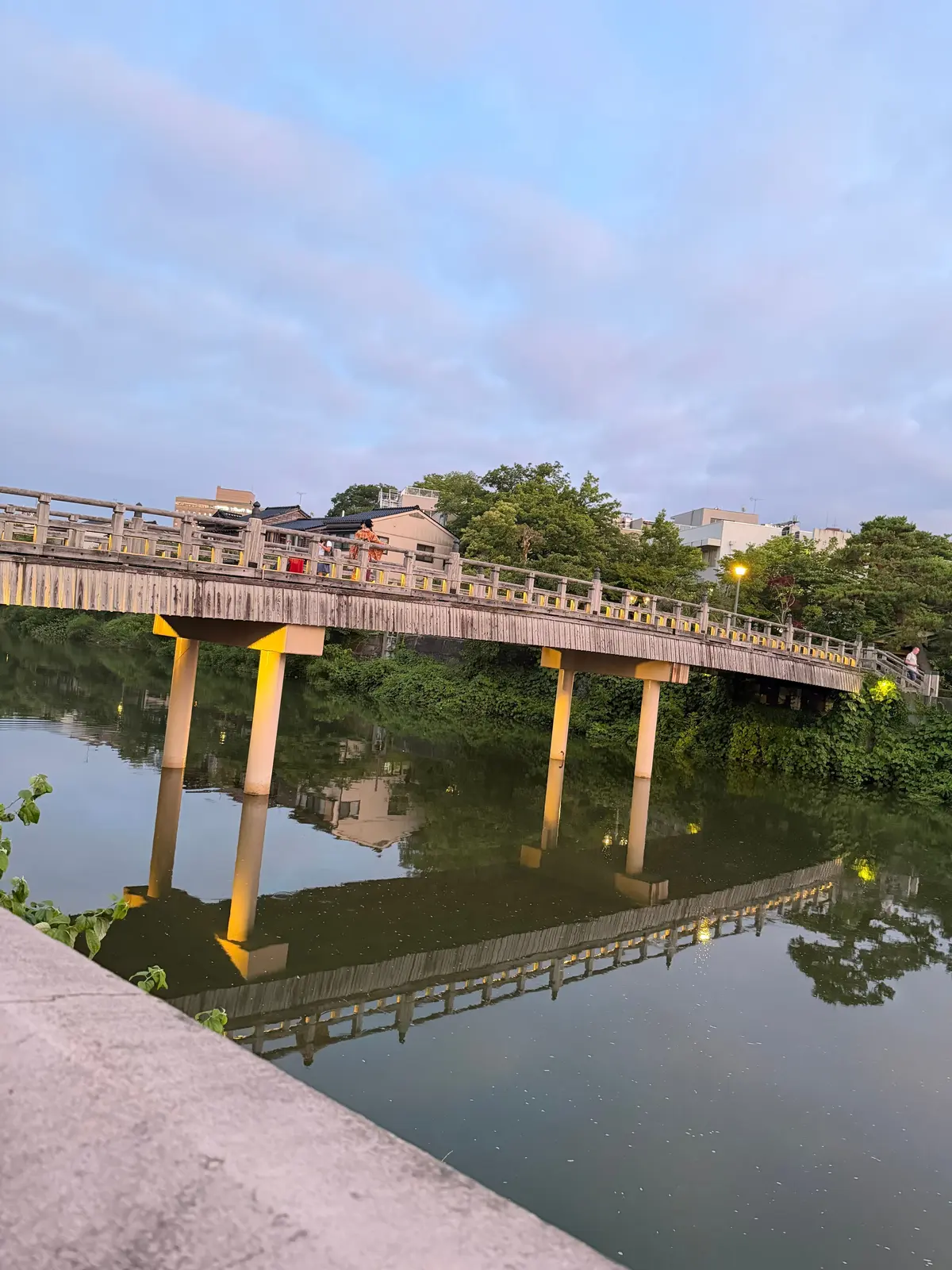 夕暮れ時、水辺に石橋が架かり、橋脚が水面に映り込んでいる。橋の上には数人が歩いている。空は淡いピンクと青色に染まっている。