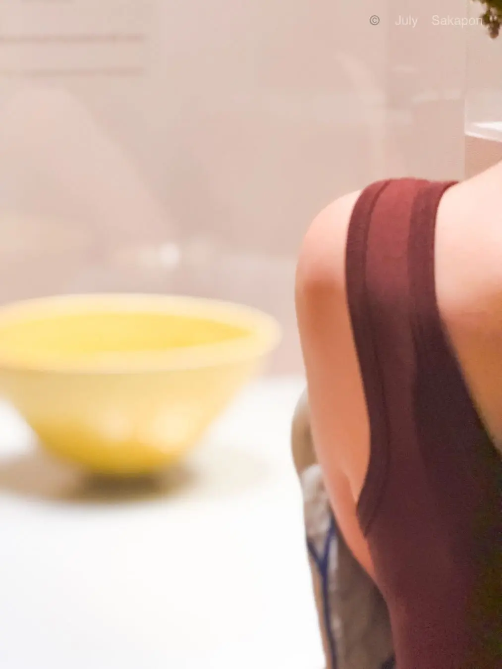 背景の白いテーブルに置かれた、黄色の丸いボウル。右側には、赤茶色の服を着た人物の肩と背中が写っている。
