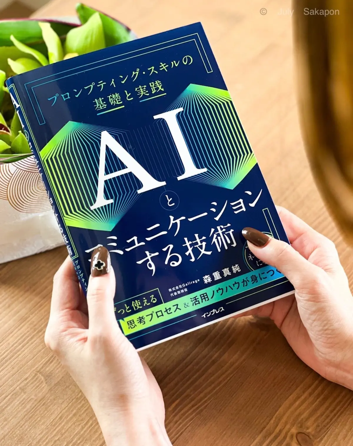 女性が、緑の葉を背景に、青と白を基調とした書籍「AIとコミュニケーションする技術」を持つ。インプレスのロゴが小さく見える。