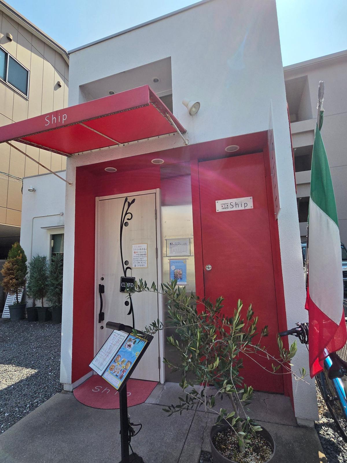 赤色の外壁に「Ship」と書かれた看板のある店舗。赤い庇があり、入り口付近にはメニュー立てと植栽がある。イタリア国旗が掲げられている。