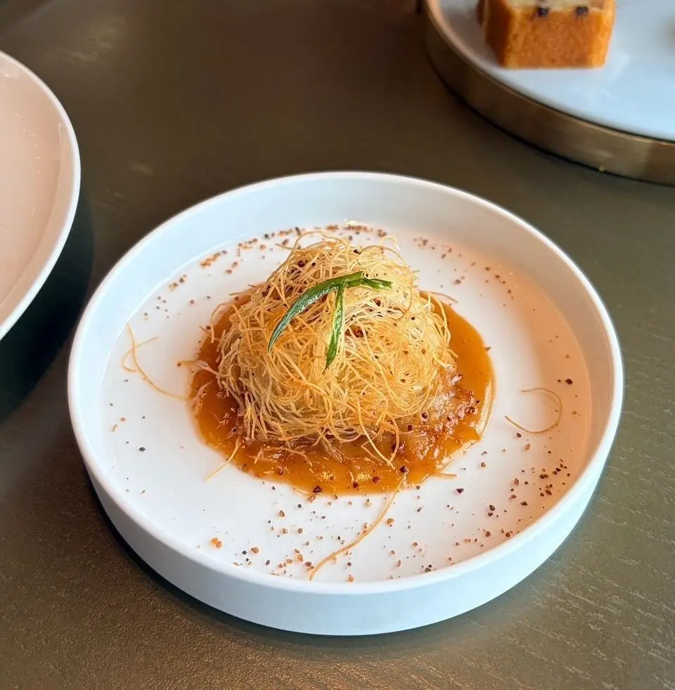 白磁の皿に盛り付けられた、オレンジ色のソースを染み込ませた細麺の料理。麺の上には緑色の草菜が添えられ、皿の周りには赤褐色の粒状のものが散らばっている。
