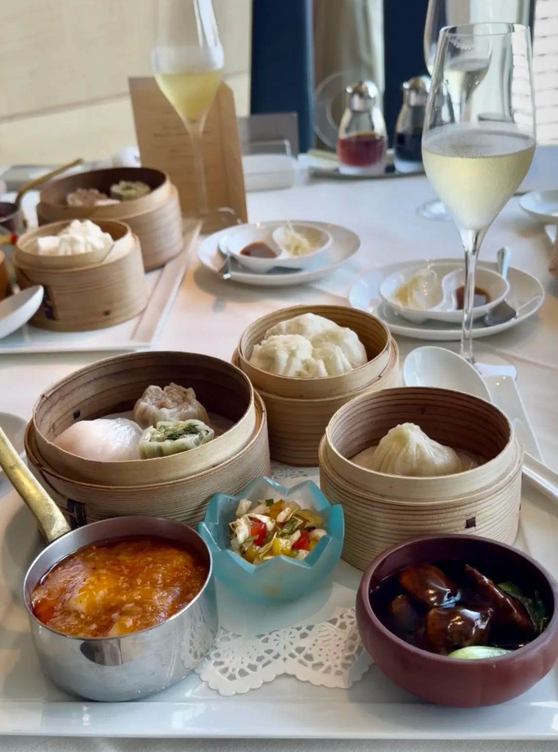杭州龍井茶（コウシュウロンジン茶）と、様々な点心（小籠包、蒸し餃子、中華まんなど）が並ぶ食卓。竹製の蒸し器に入った点心、ソース、薬味、調味料が盛り付けられている。点心師がつくる繊細な点心の数々と王道メニュー。
