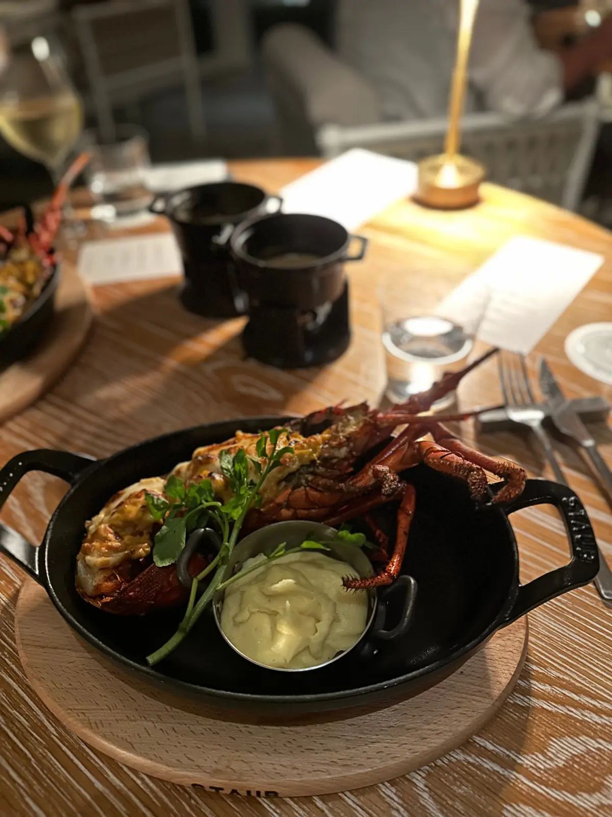 黒い Staub 製の鍋に入った、調理されたロブスター。ソースとハーブが添えられ、木製のトレイに乗っている。背景にはテーブル、グラス、照明が写る。