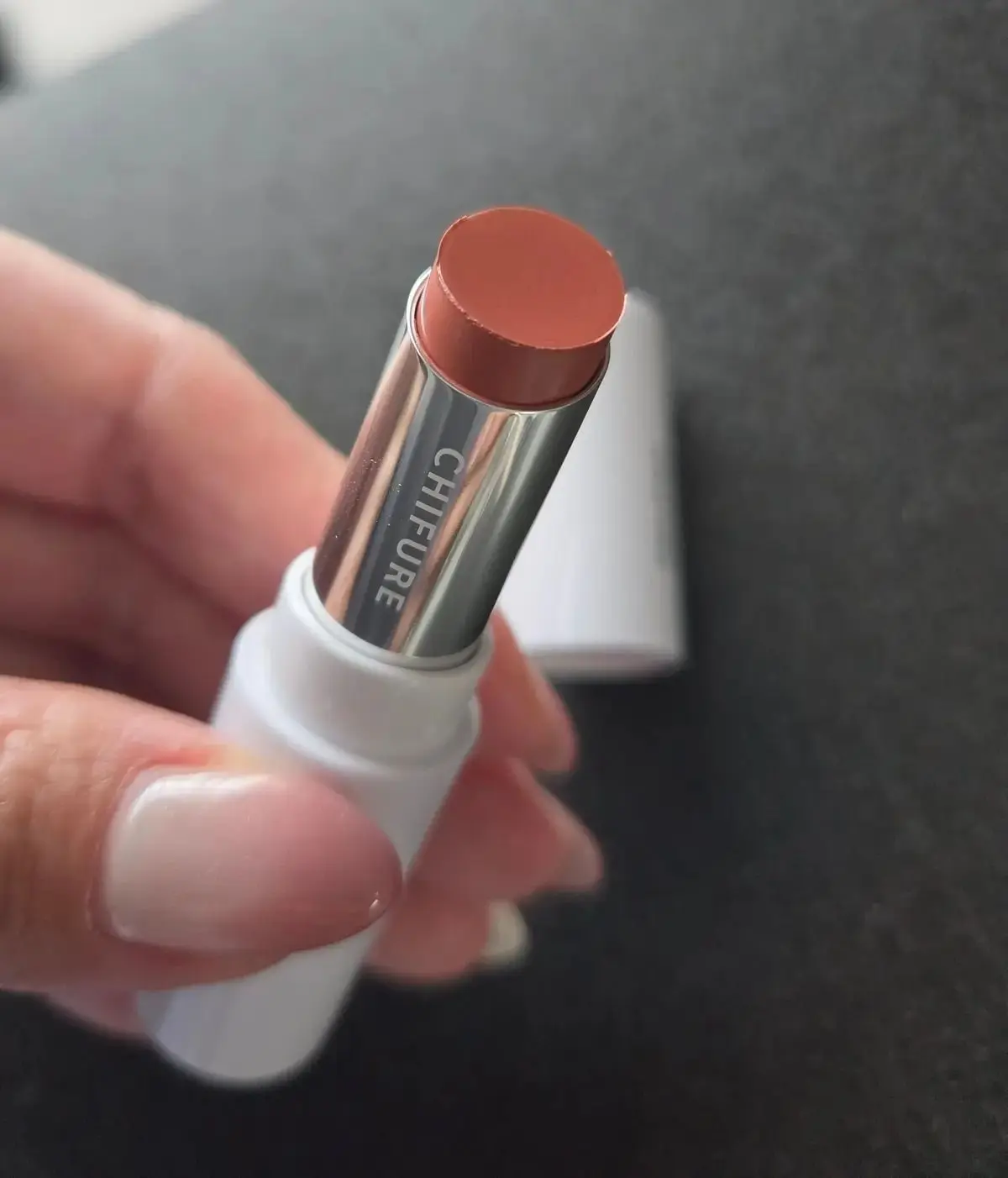 手のひらで支えられた、CHIFURE（チフューレ）のリップスティック。オレンジがかったベージュ色のスティックで、シルバーのリングと白いケースが付いている。