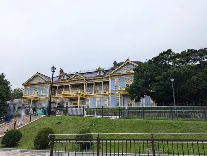 青と黄色のコントラストが美しい、明治時代の洋館。緑の芝生の上に建ち、階段と黒いフェンスが見える。背景には緑豊かな丘が広がっている。