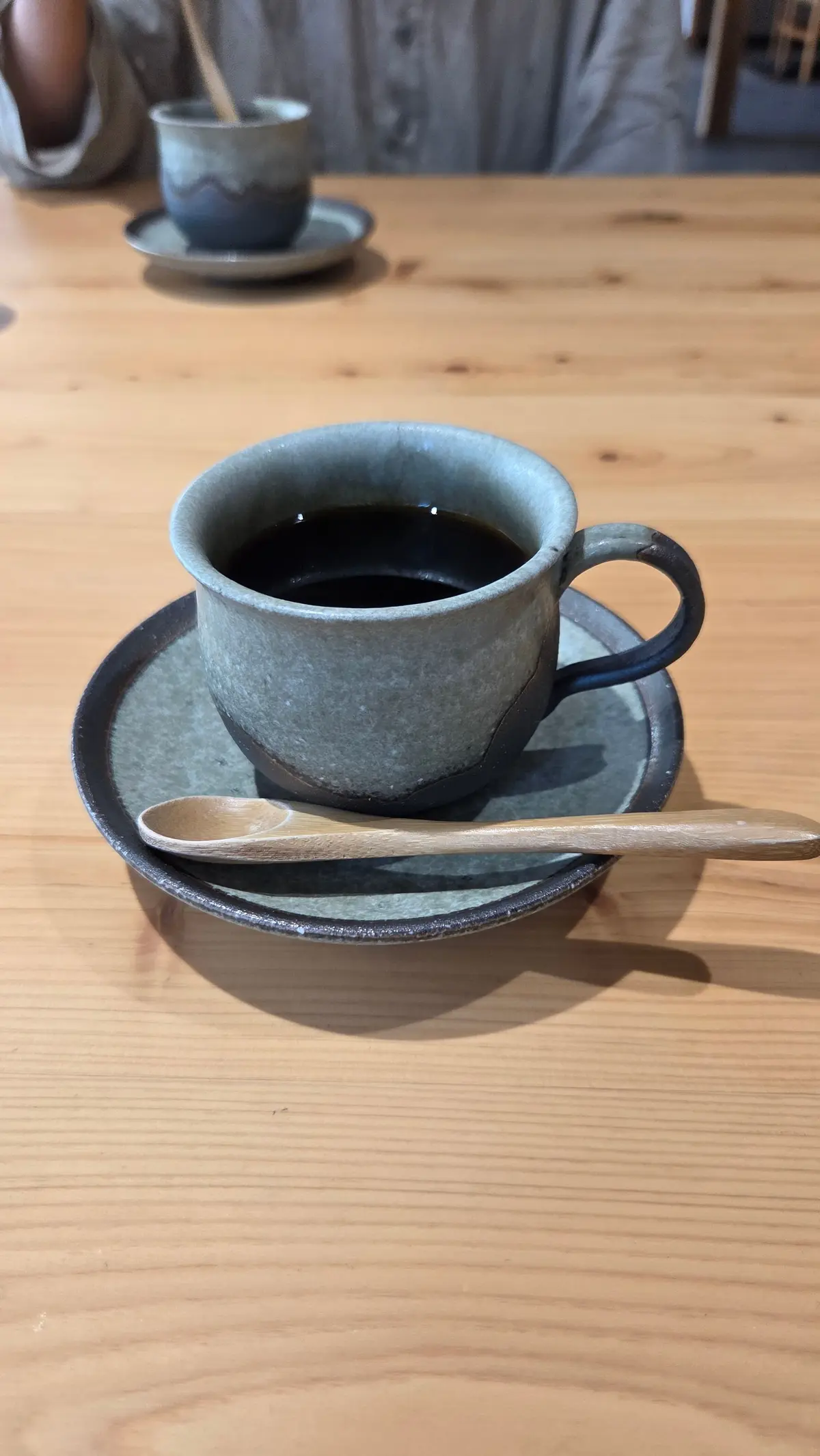 木製のテーブルに置かれた、灰色の素朴な陶器のコーヒーカップとソーサー。カップには黒いコーヒーが注がれ、横には木のスプーンが置かれている。背景には、別のカップとソーサーが見える。