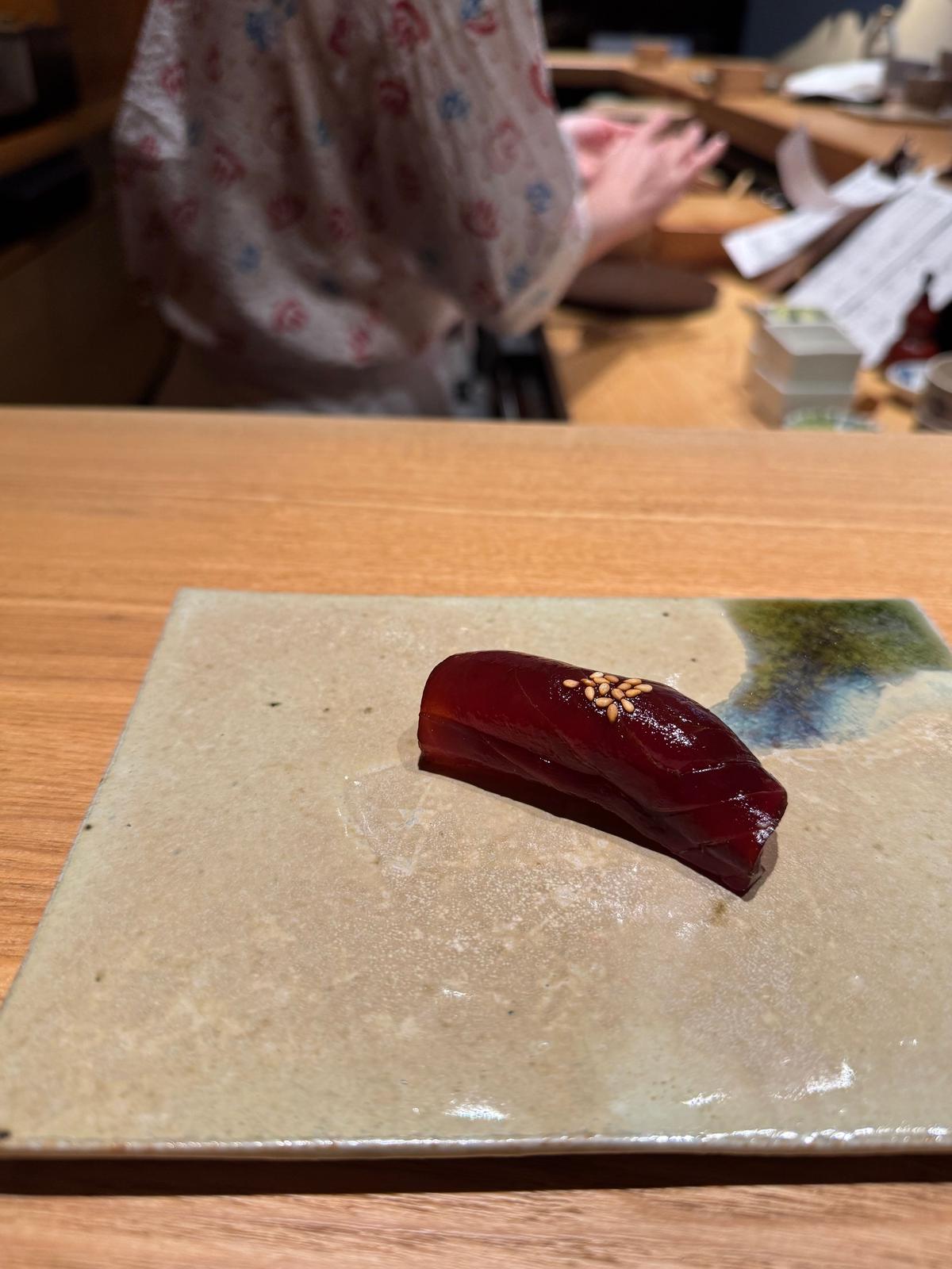 茶色の長方形の皿に、赤色のマグロの刺身が乗っている。刺身の上には、金色の粒状のものが散らされており、その横には緑色のアクセントがある。