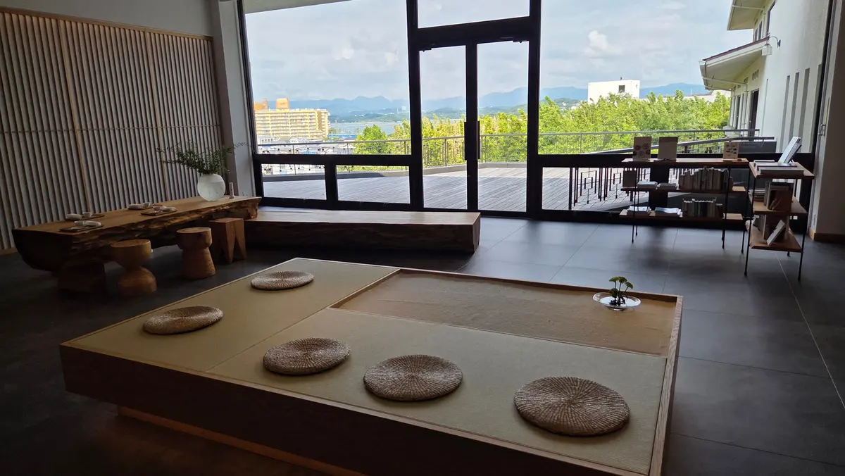 広い部屋に、中央に畳のローテーブル、左に木製の長テーブルと椅子、右にはテラスへの大きな窓がある。窓の外には海と山が見える。