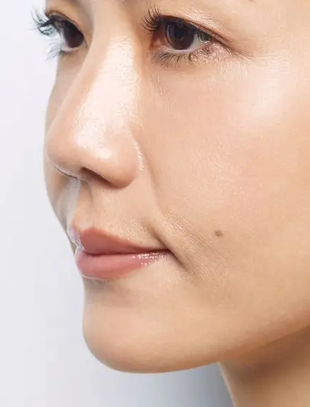 白い背景をバックにした女性の横顔。黒髪を後ろでまとめ、シンプルなベージュのトップスを着用。自然な表情で、肌はきめ細かく、右頬に小さなそばかすがある。
