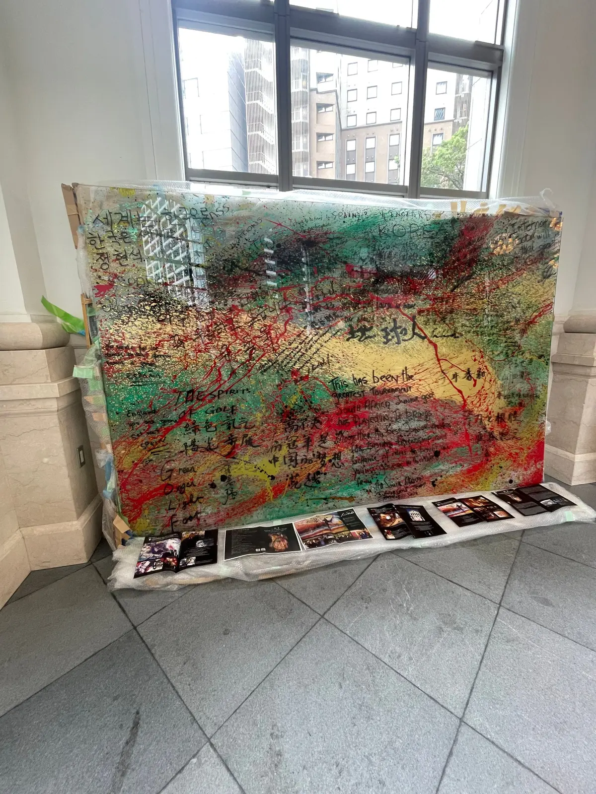抽象的な絵画が、窓の前に置かれている。緑、赤、黄色の筆致が混ざり、手書きの文字が散りばめられている。絵画の下には、複数のポスターが並んでいる。