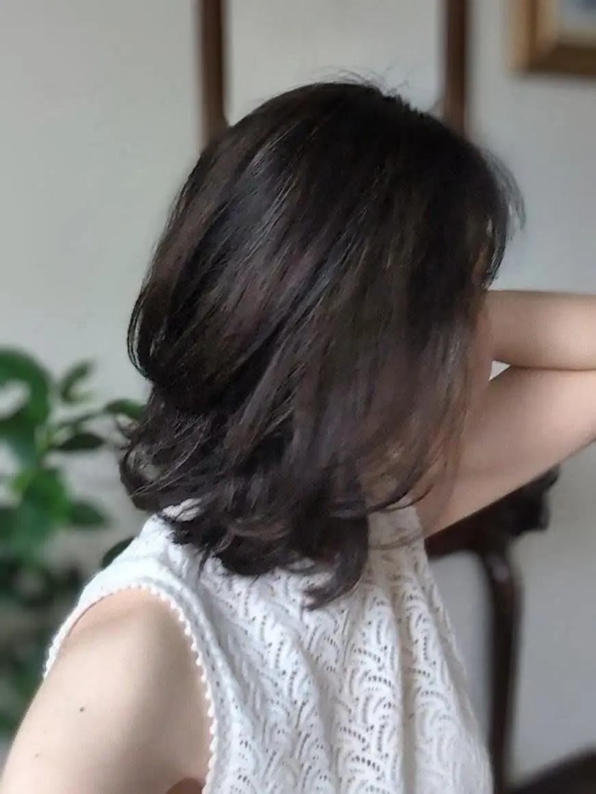 肩くらいの長さのダークブラウンの髪を後ろで束ねた女性。白いレースのトップスを着ており、背景には植物と木製のフレームが見える。