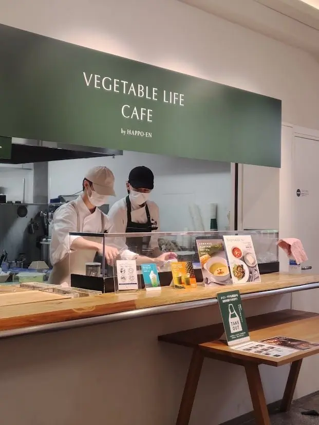 カフェのカウンターで、白衣を着た男性店員がマスクを着用し、調理器具を使って作業している様子。背景には「VEGETABLE LIFE CAFE by HAPPO-EN」のロゴがある。