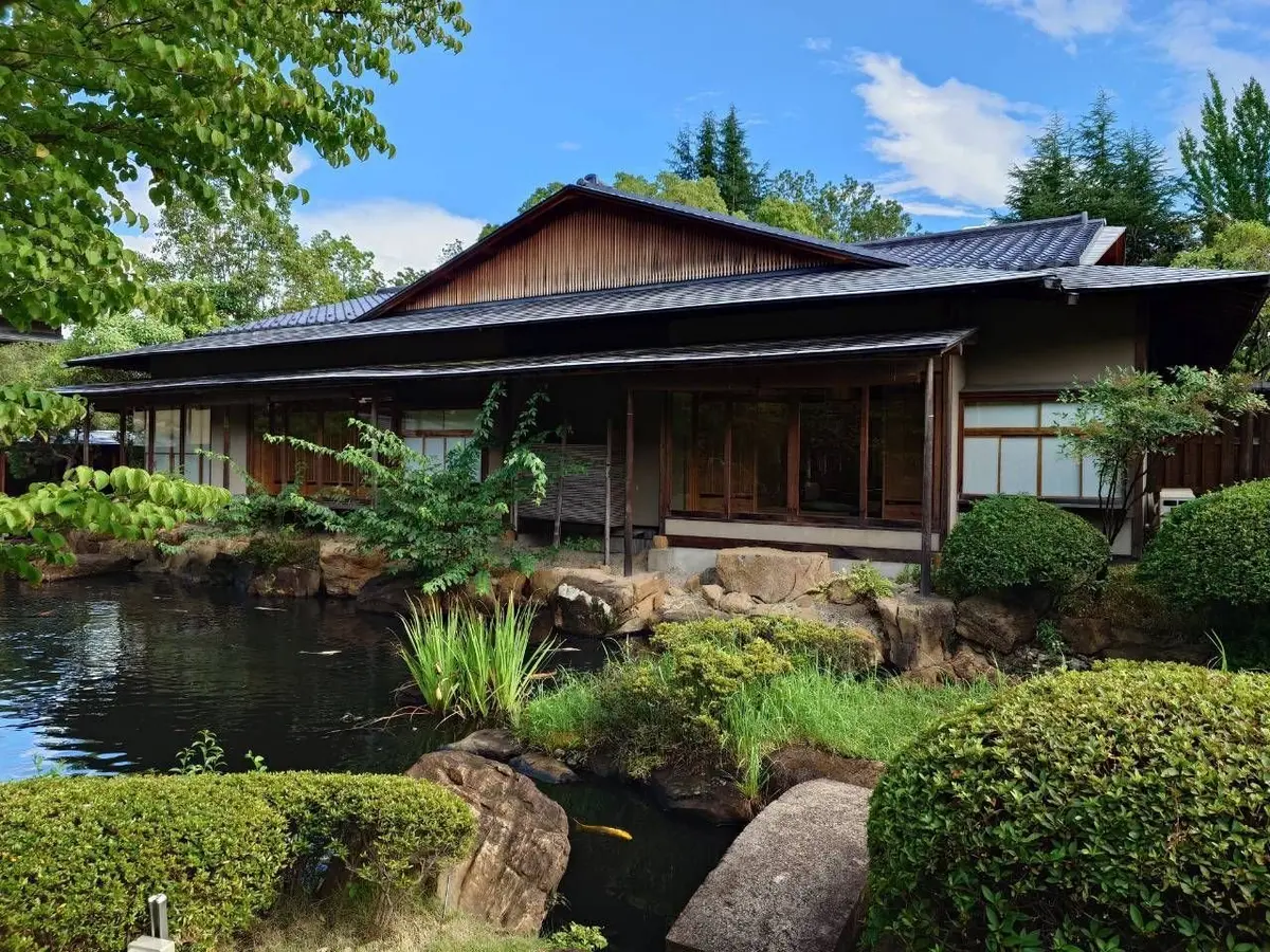 池のほとりに佇む、木造の日本家屋。瓦屋根、縁側、大きな窓が特徴的。手前に苔むした岩や緑の植物。常磐ホテルの離れ「九重」