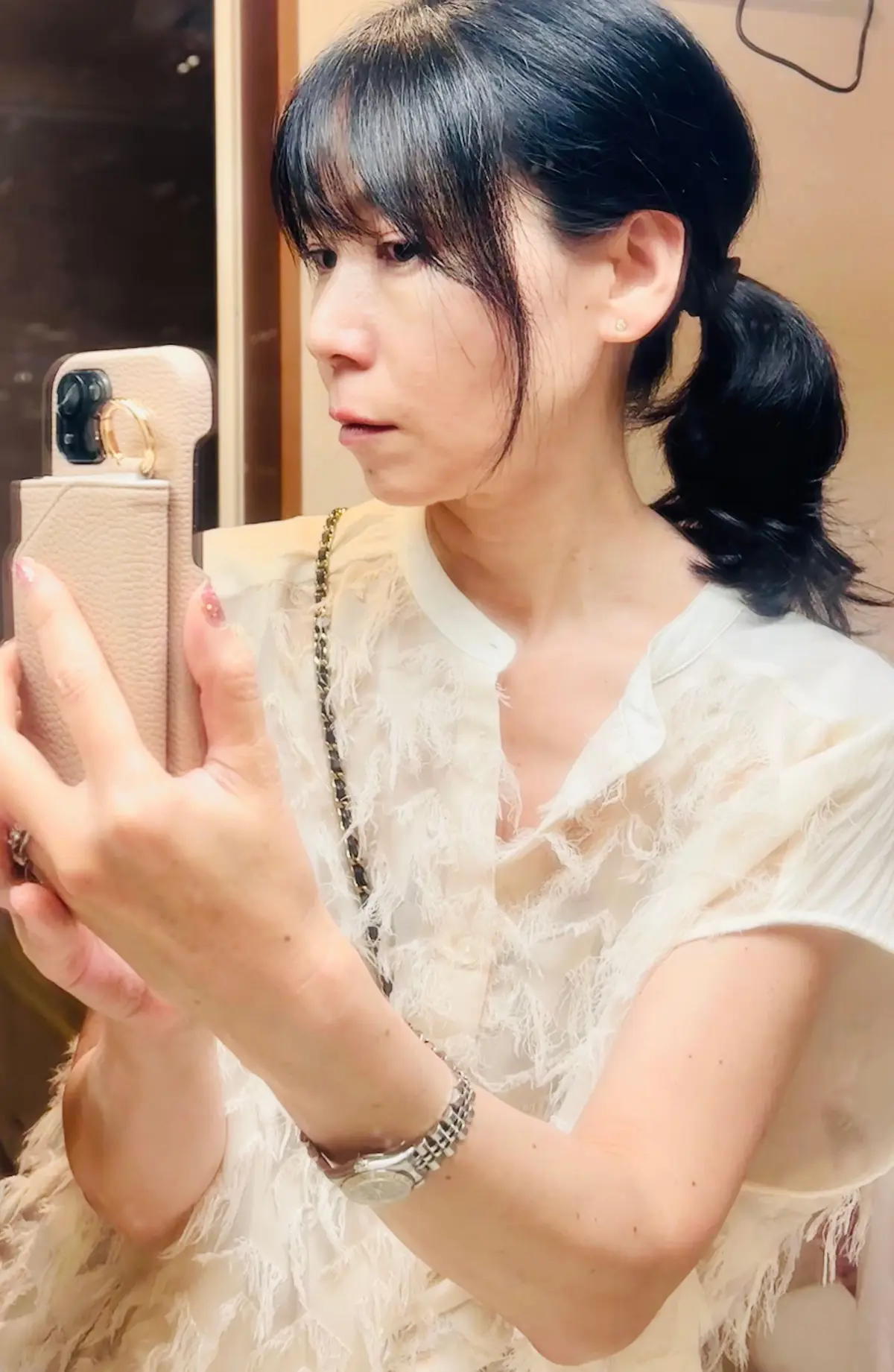 白いフリル付きトップスを着た女性が、自撮り写真でこちらを見つめている。黒髪のボブに、低い位置でポニーテール。右手にはディオールのバッグを持ち、左手首には腕時計をしている。背景は木目調の壁。